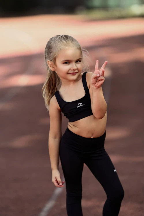 Mała modelka stoi na bieżni, widok z przodu, czarne legginsy dziewczęce z przeszyciami ActiveGlow.