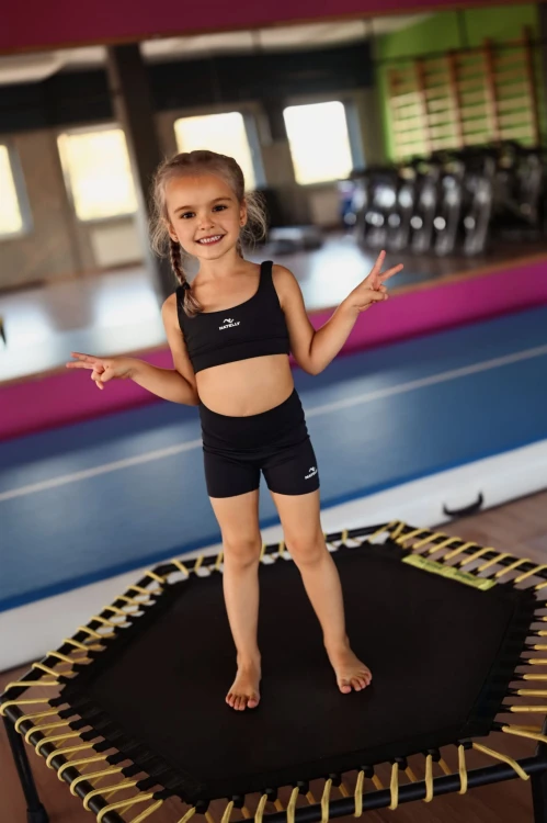 Mała modelka stoi na trampolinie w czarnych krótkich getrach dla dzieci